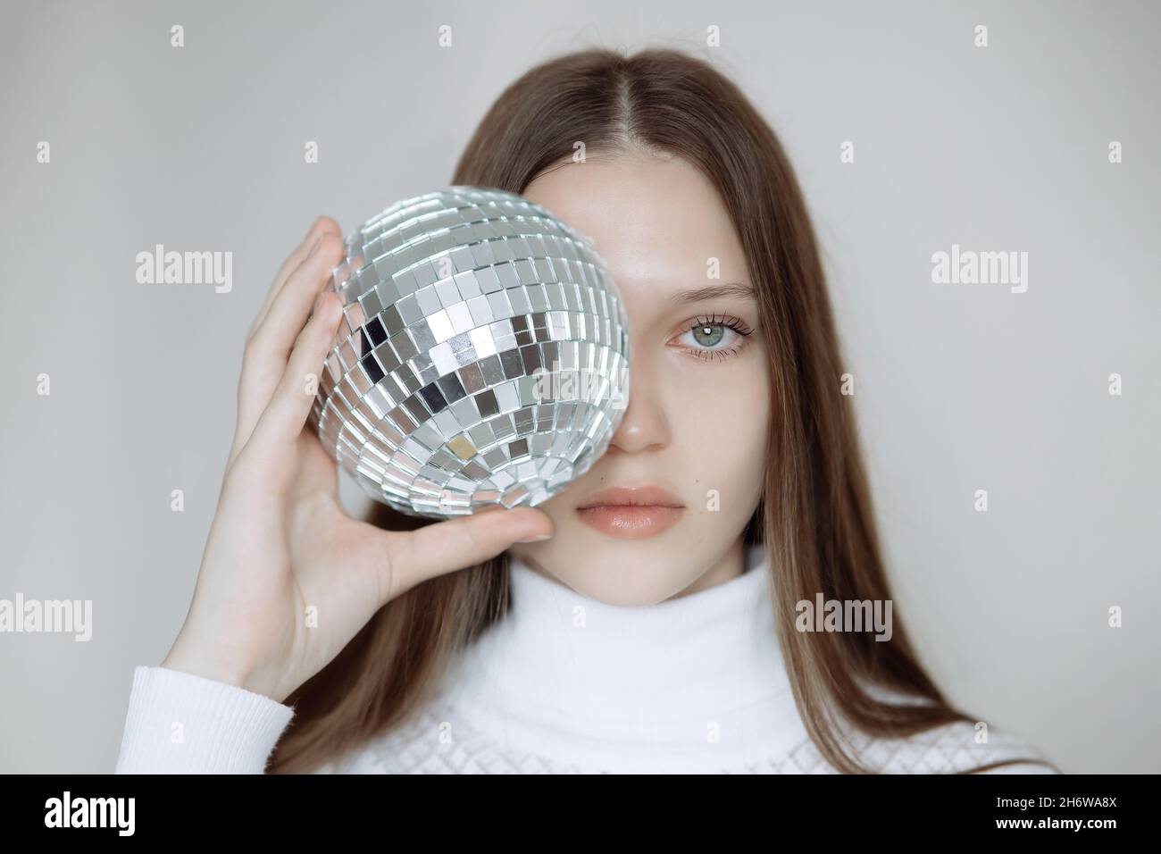 Retrato de moda de una adolescente con una bola de disco, cubriendo su  cara. Una chica dulce con ojos expresivos y un jersey blanco sostiene una  esfera de espejo Fotografía de stock -