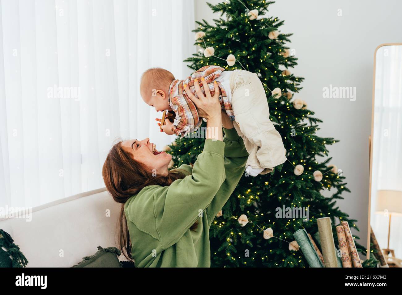 Una joven madre de pelo rojo juega y se divierte con su bebé, levantando a su hija por encima de su cabeza. Navidad familiar en casa. Foto de stock