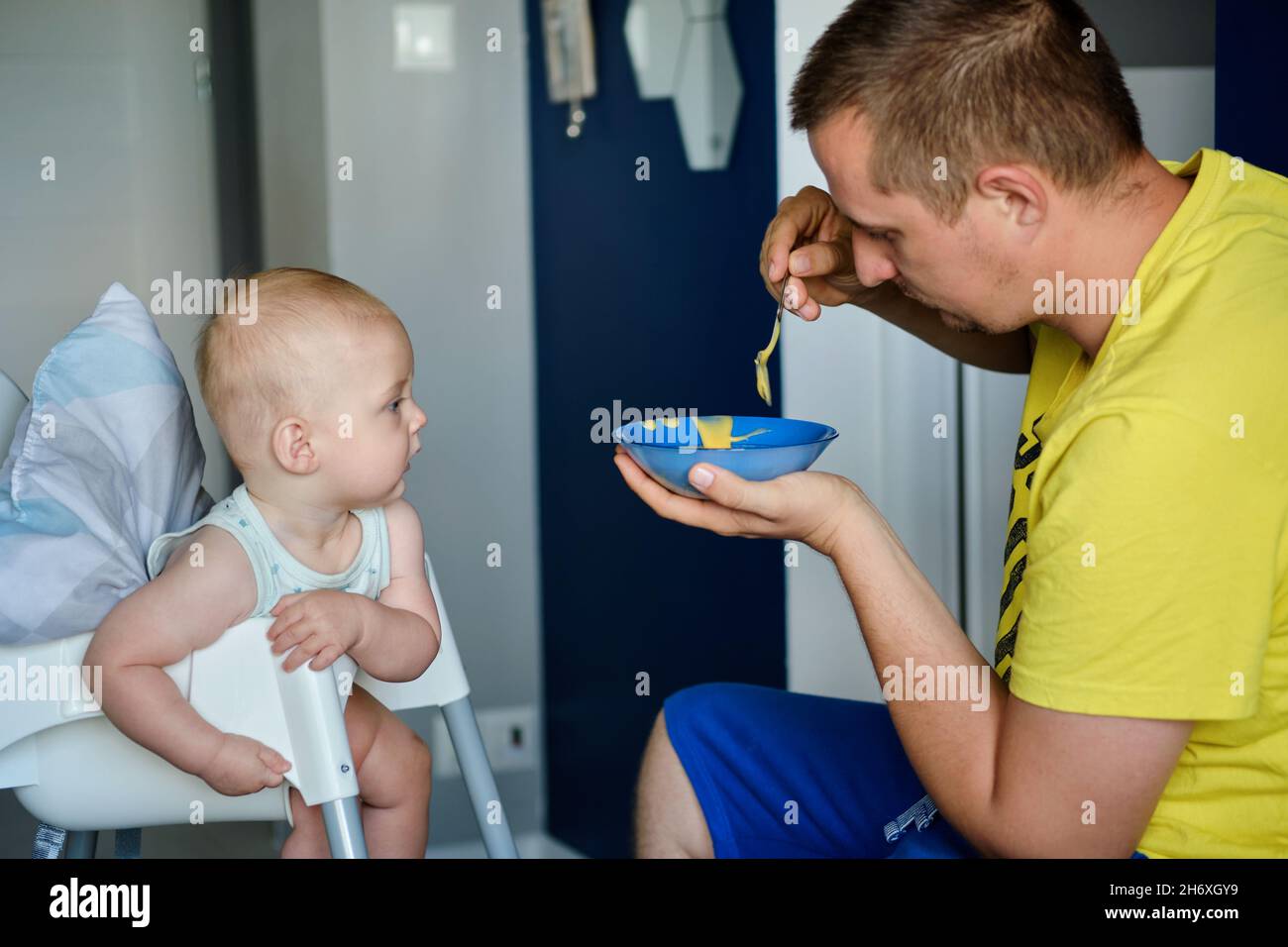 Un padre joven alimenta a un bebé lindo con comida amarilla para bebés Foto de stock