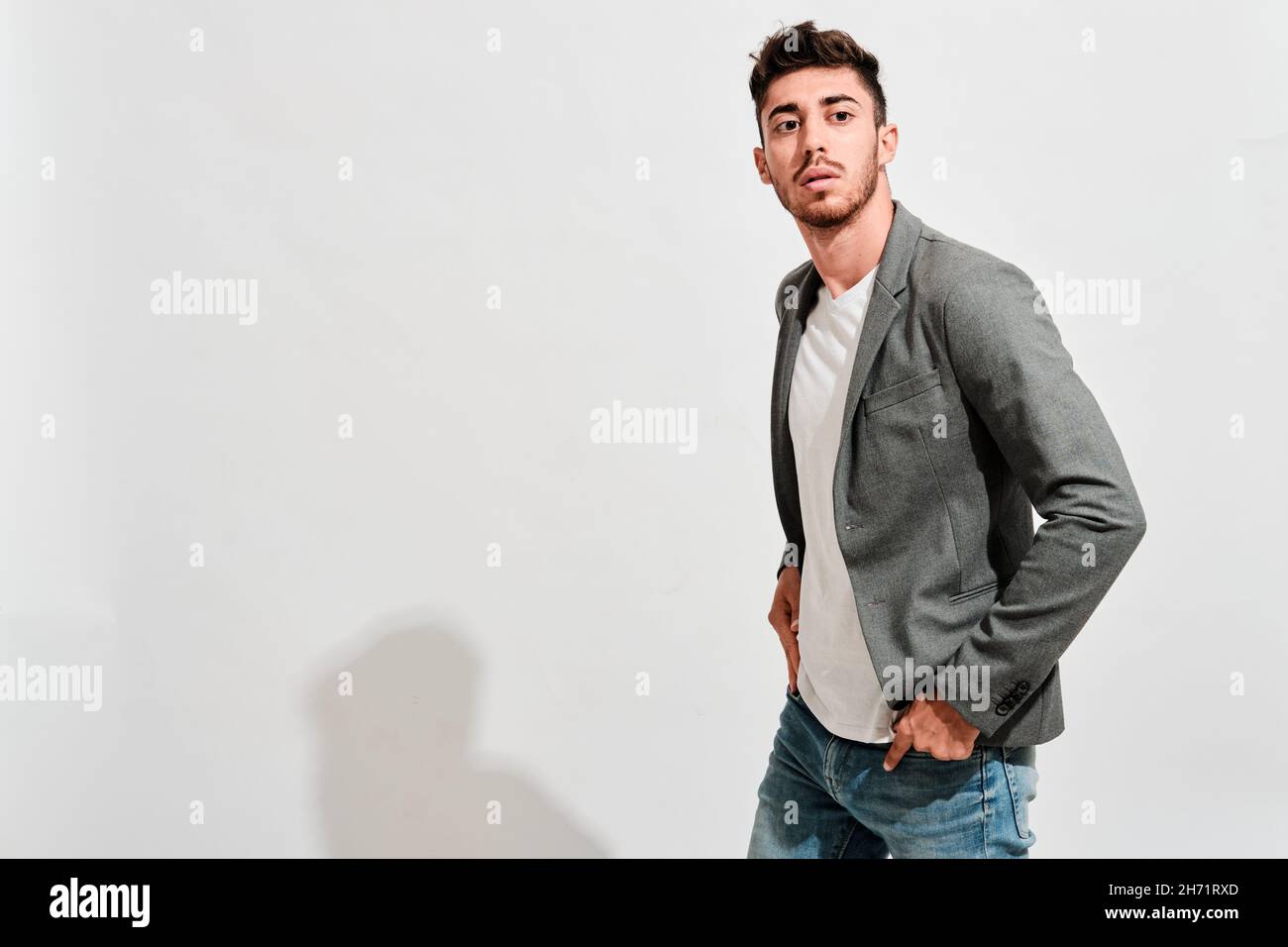 Hombre joven con jeans, un tshit blanco y un blazer gris caminando delante  de una pared blanca con sus manos dentro de los bolsillos de los jeans  Fotografía de stock - Alamy
