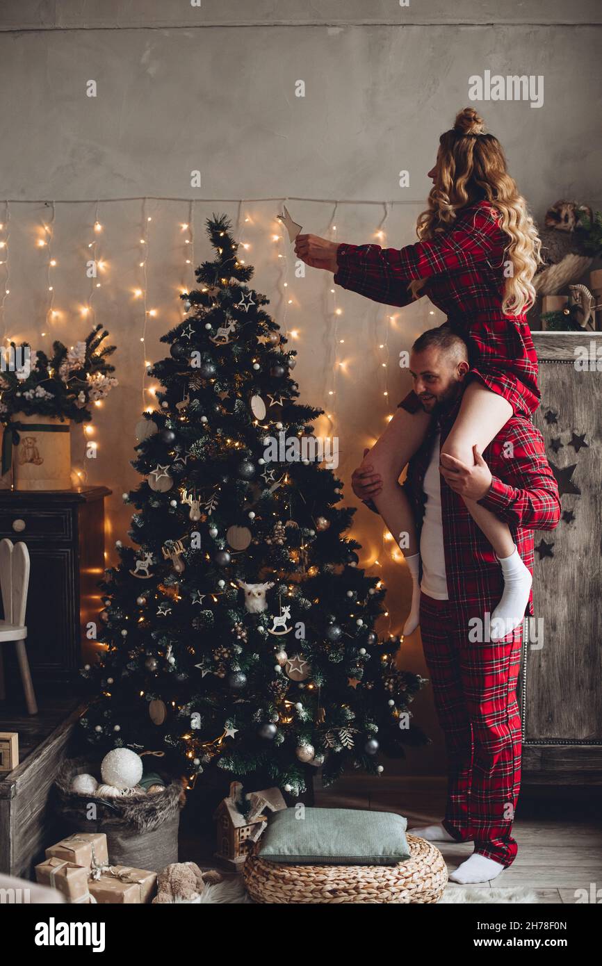Pareja amorosa en pijama rojo de cuadros que adorna el árbol de Navidad.  Mujer sentada sobre los hombros del hombre Fotografía de stock - Alamy