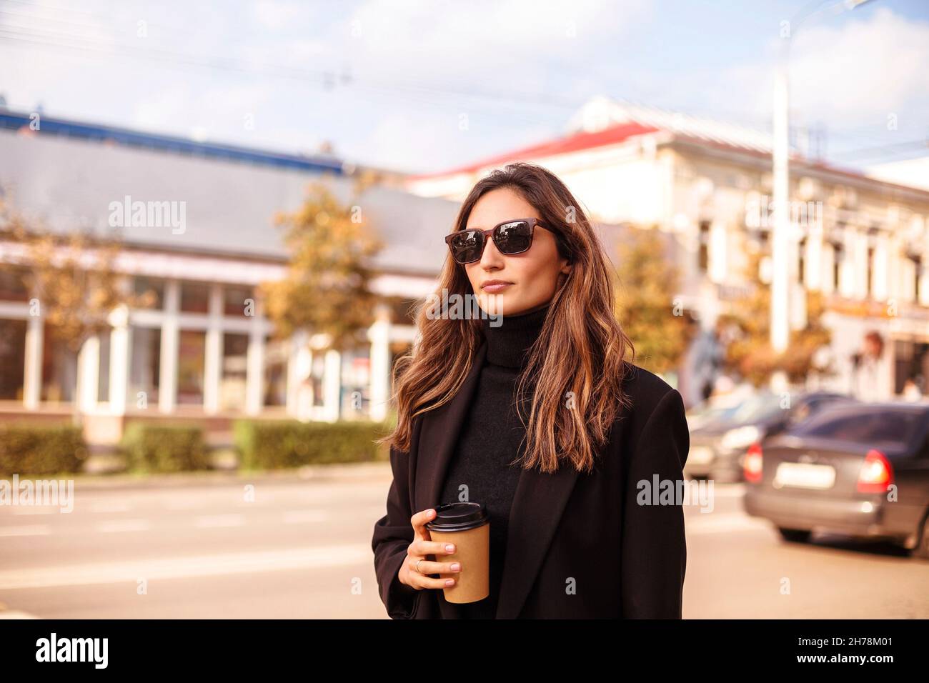 Joven elegante mujer con estilo en ropa negra y gafas de sol que sostienen  una taza de café para llevar mientras camina por la calle de la ciudad,  hermosa señora de negocios