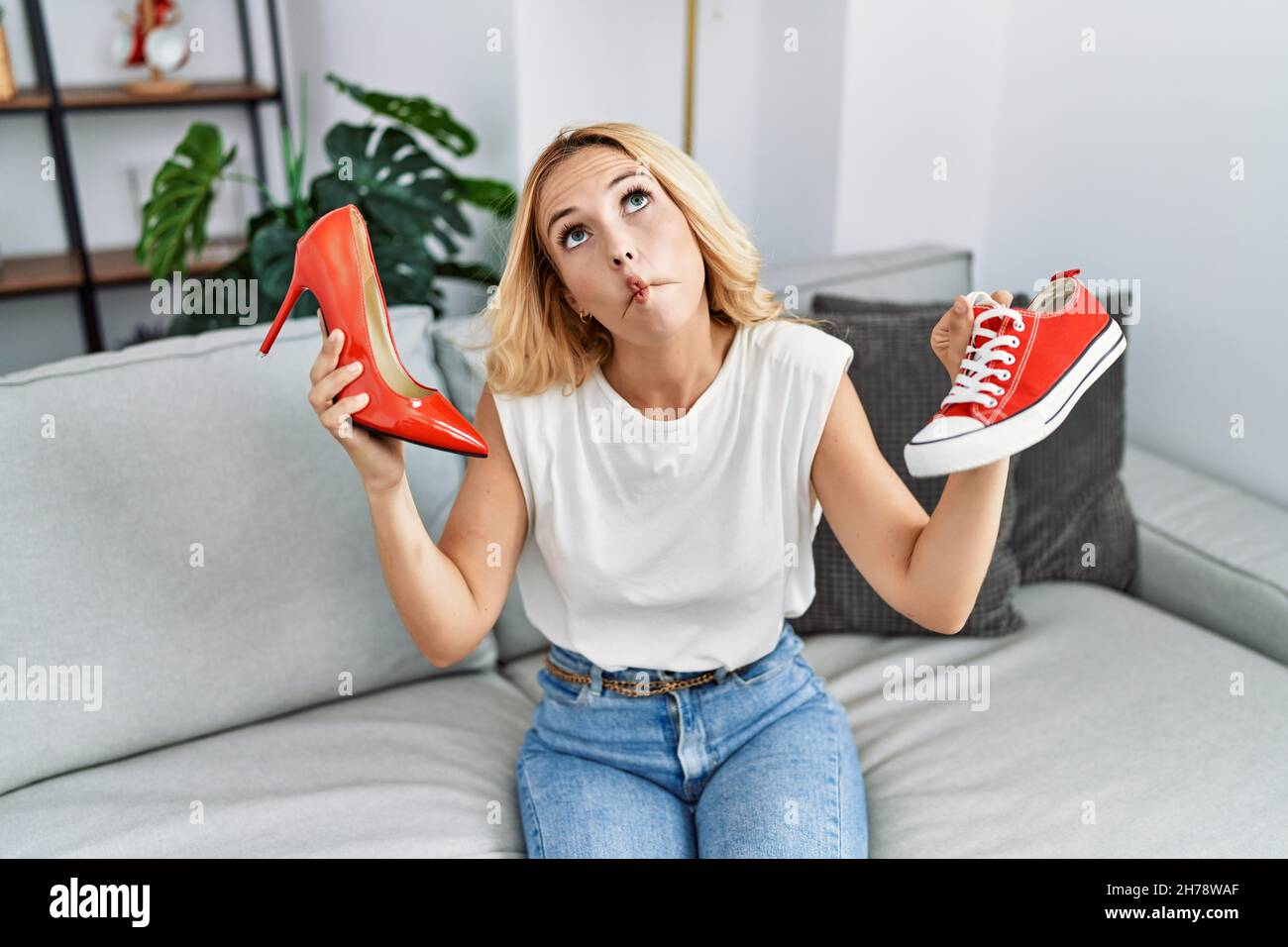 Hermosa mujer rubia elegir zapatos de tacón alto zapatillas de deporte en casa haciendo la cara de los peces con la boca y ojos entrecerrados, loco y cómico Fotografía de stock -
