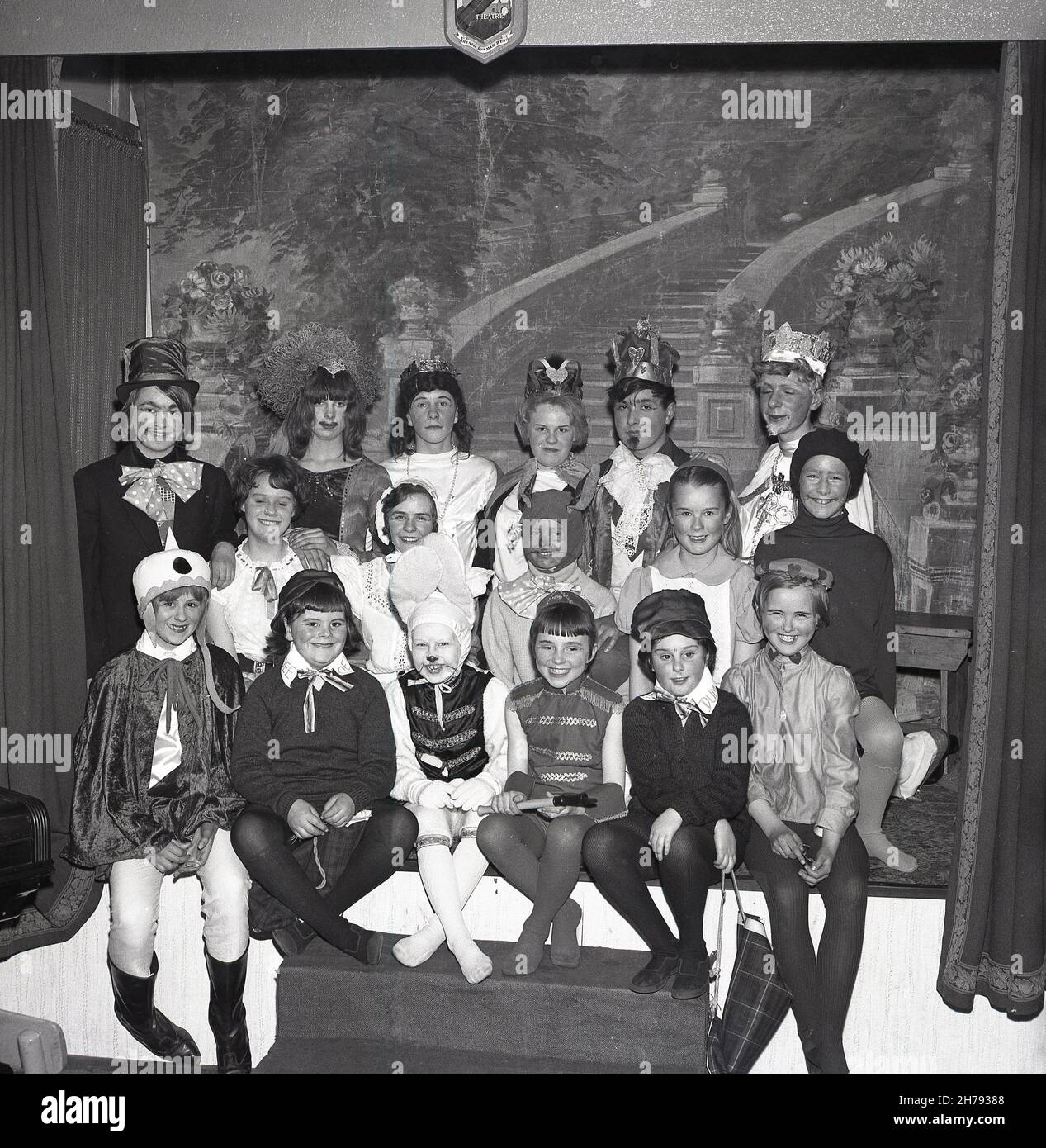1965, histórico, sentados juntos en un pequeño escenario, grandes sonrisas de un grupo de niños de un centro juvenil vestidos con sus trajes para la obra, Alice in Wonderland, Fife, Escocia, Reino Unido. Foto de stock