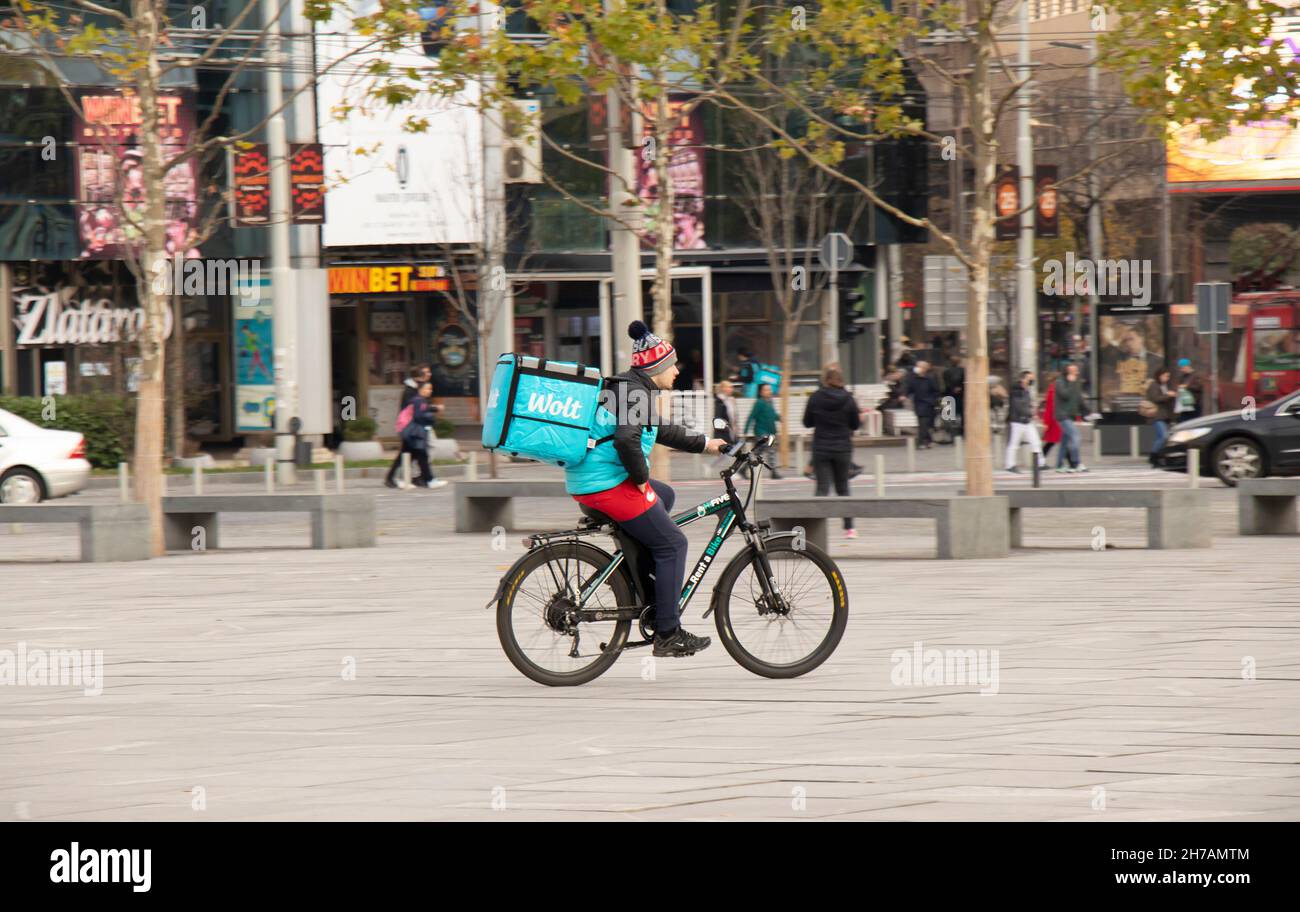 Belgrado, Serbia - 17 de noviembre de 2021: Persona que trabaja para el servicio de entrega de alimentos de la ciudad de Wolt en bicicleta a través de la plaza de la ciudad Foto de stock