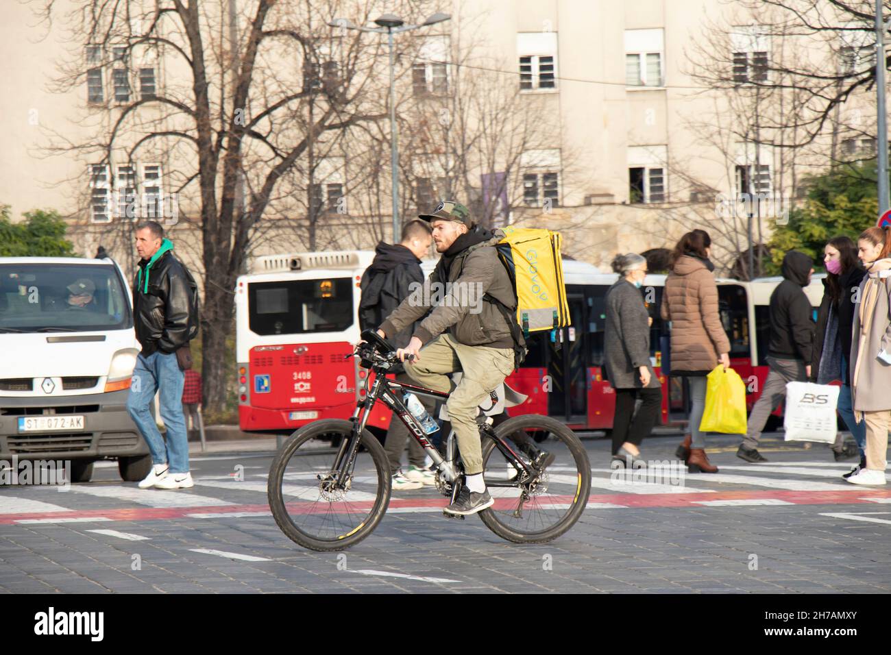 Belgrado, Serbia - 17 de noviembre de 2021: Persona que trabaja para el servicio de entrega de alimentos de la ciudad de Glovo en bicicleta mientras cruza la ajetreada calle de la ciudad Foto de stock