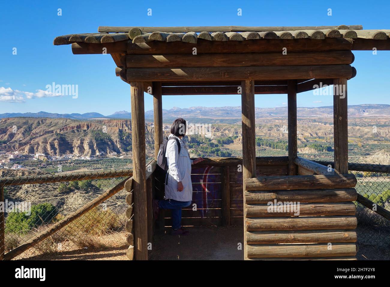 Mujer de 50 años de edad con chubasquero y mochila admirando el paisaje de los badlands de Marchal (España) y sus casas cueva desde un mirador de madera Foto de stock