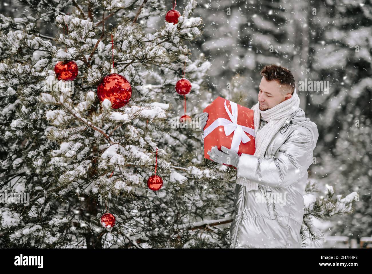 El joven barbudo, con un elegante abrigo plateado, está en el árbol de Navidad decorado con una caja de regalo de vacaciones al aire libre Foto de stock