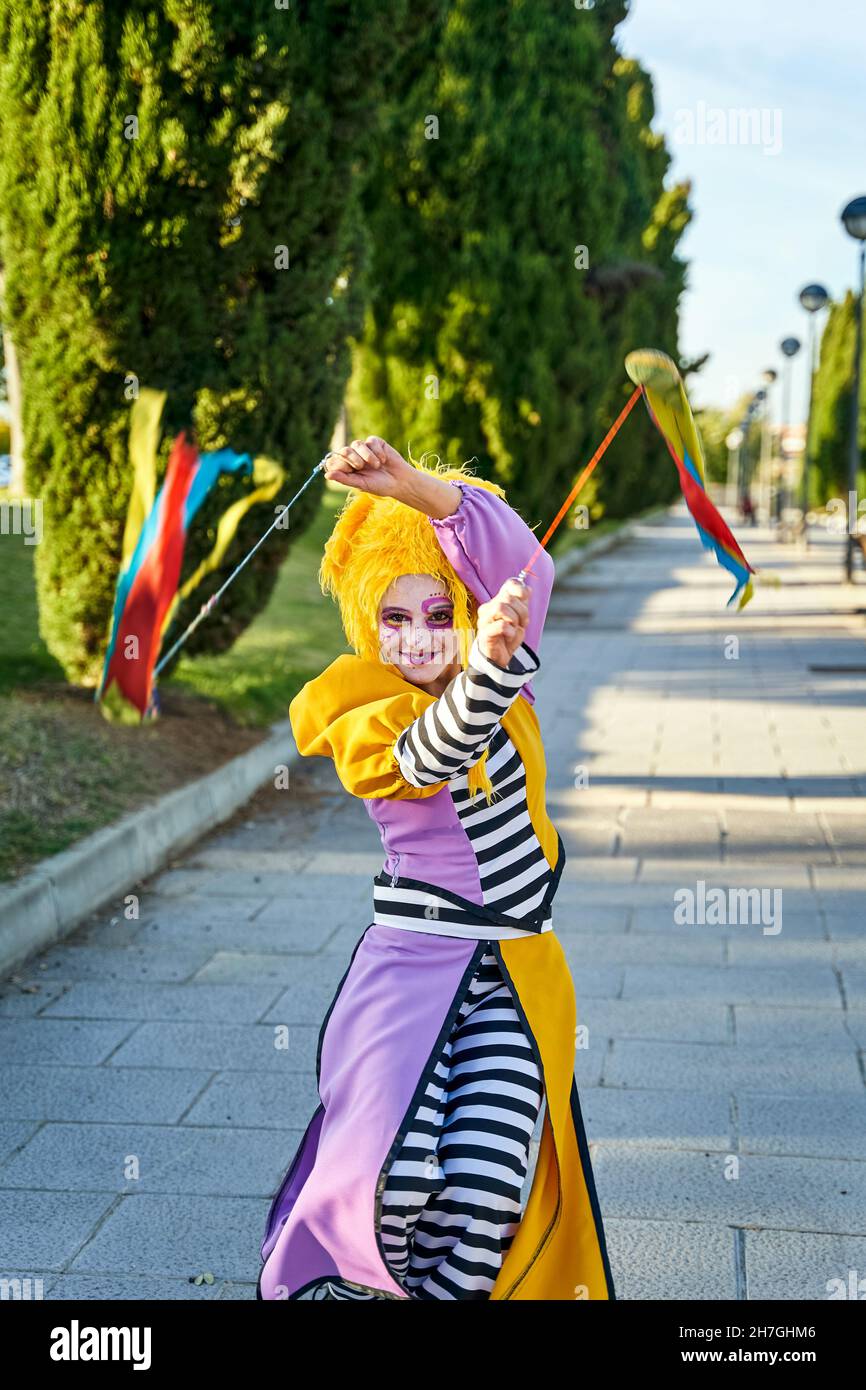 Feliz payaso femenino con rostro pintado con divertido traje y peluca amarilla, sonriendo y caminando por el camino en el parque con coloridos poi en las manos durante el desfile Foto de stock