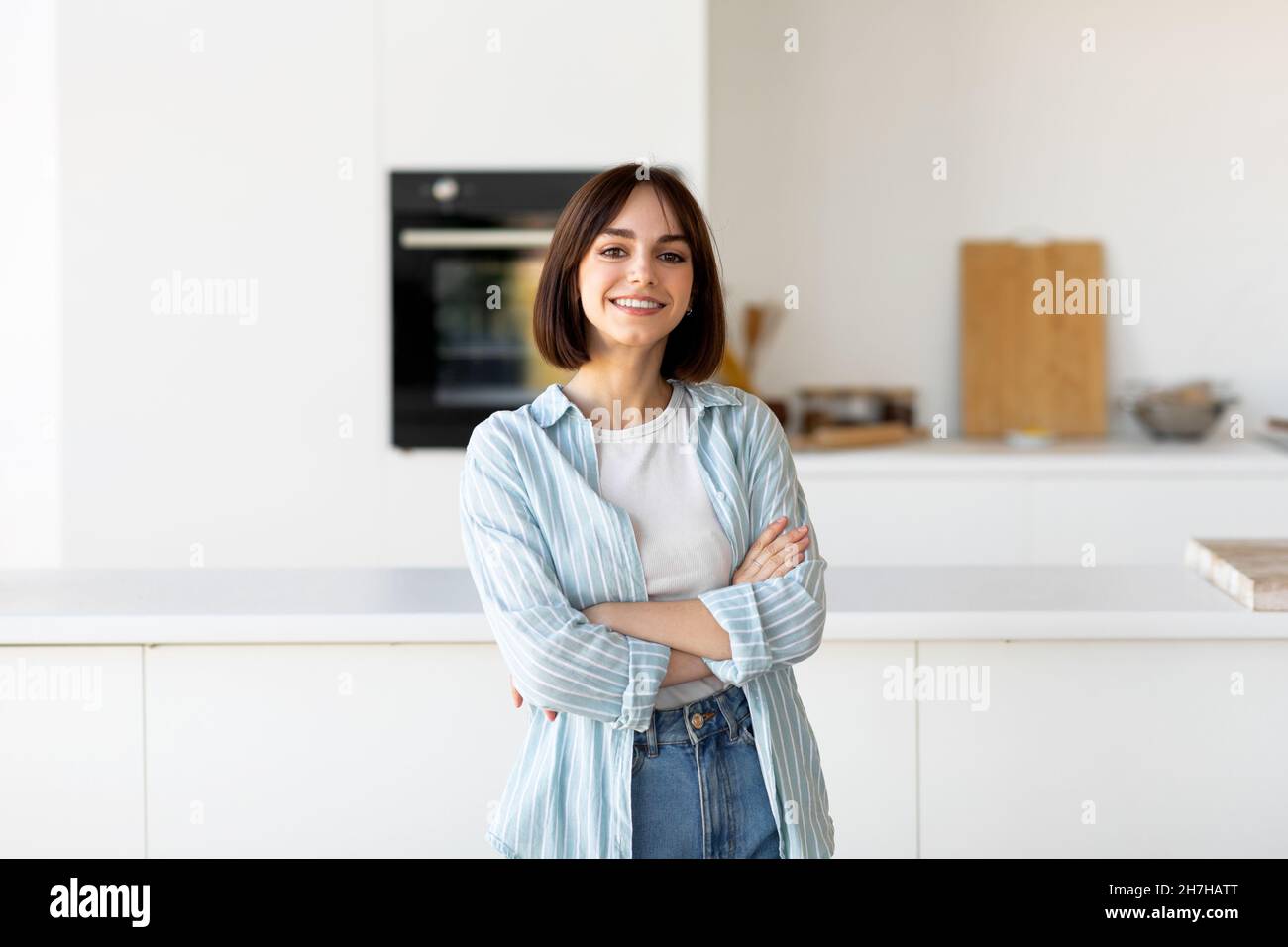 Feliz persona. Retrato de joven posando con las manos cruzadas, sonriendo a la cámara, de pie en el interior de la cocina moderna Foto de stock