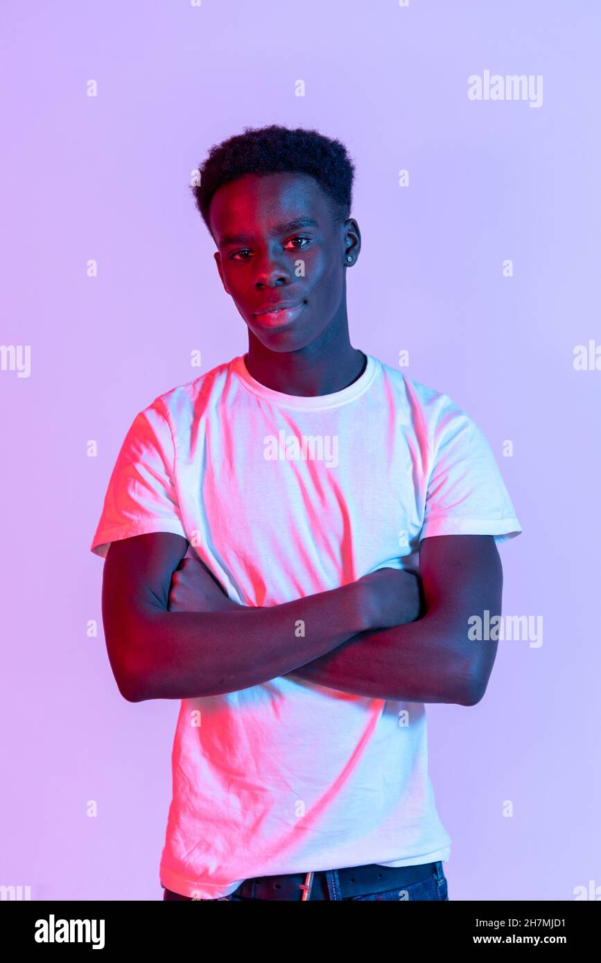 Modelo masculino joven afroamericano seguro con pelo oscuro en camiseta blanca de pie contra fondo púrpura con brazos cruzados y mirando a la leva Foto de stock