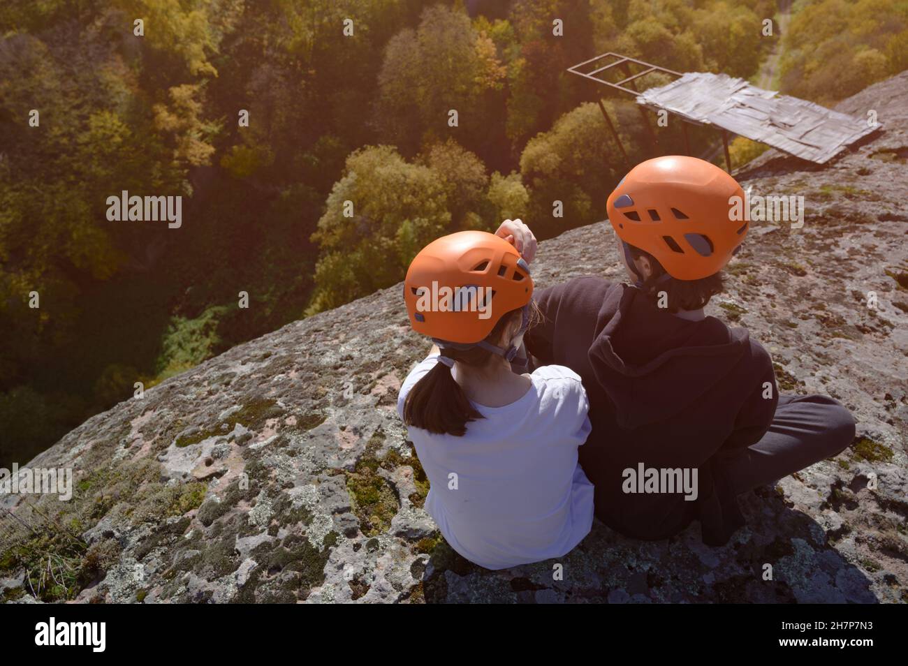 dos niños pequeños en casco naranja sentados en la cima de la alta montaña rocosa con el bosque abajo Foto de stock