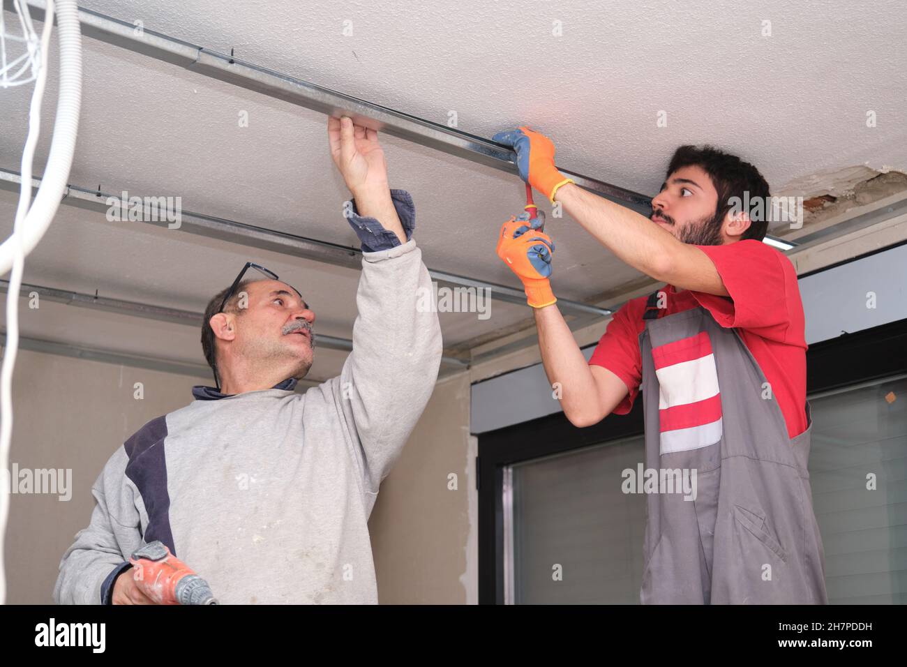 Dos trabajadores que ensamblan un marco de metal de techo suspendido con un destornillador. Foto de stock