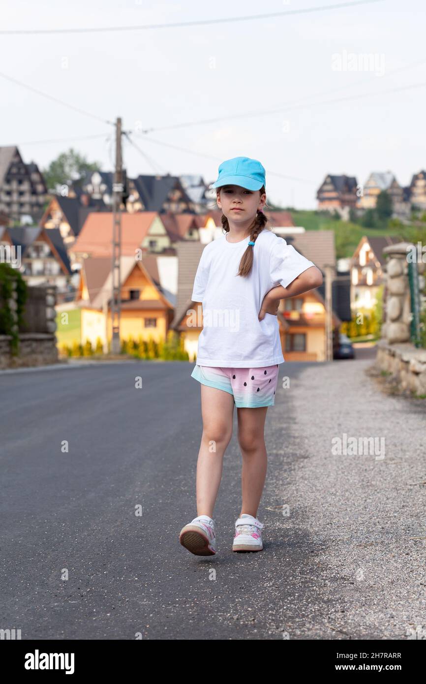 Niño en edad elemental, niña en edad escolar caminando solo por un pueblo pequeño, dando un paseo, dando un paseo por la calle, en el área rural, una persona, camiseta blanca en blanco, bajo Foto de stock