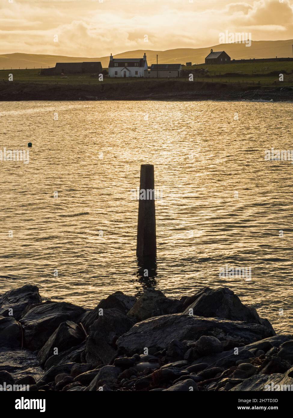 Vista al amanecer al otro lado de la bahía desde Melby Pier, Melby, cerca de Sandness, con el poste que marca el extremo de la slipway en el primer plano, Shetland Foto de stock