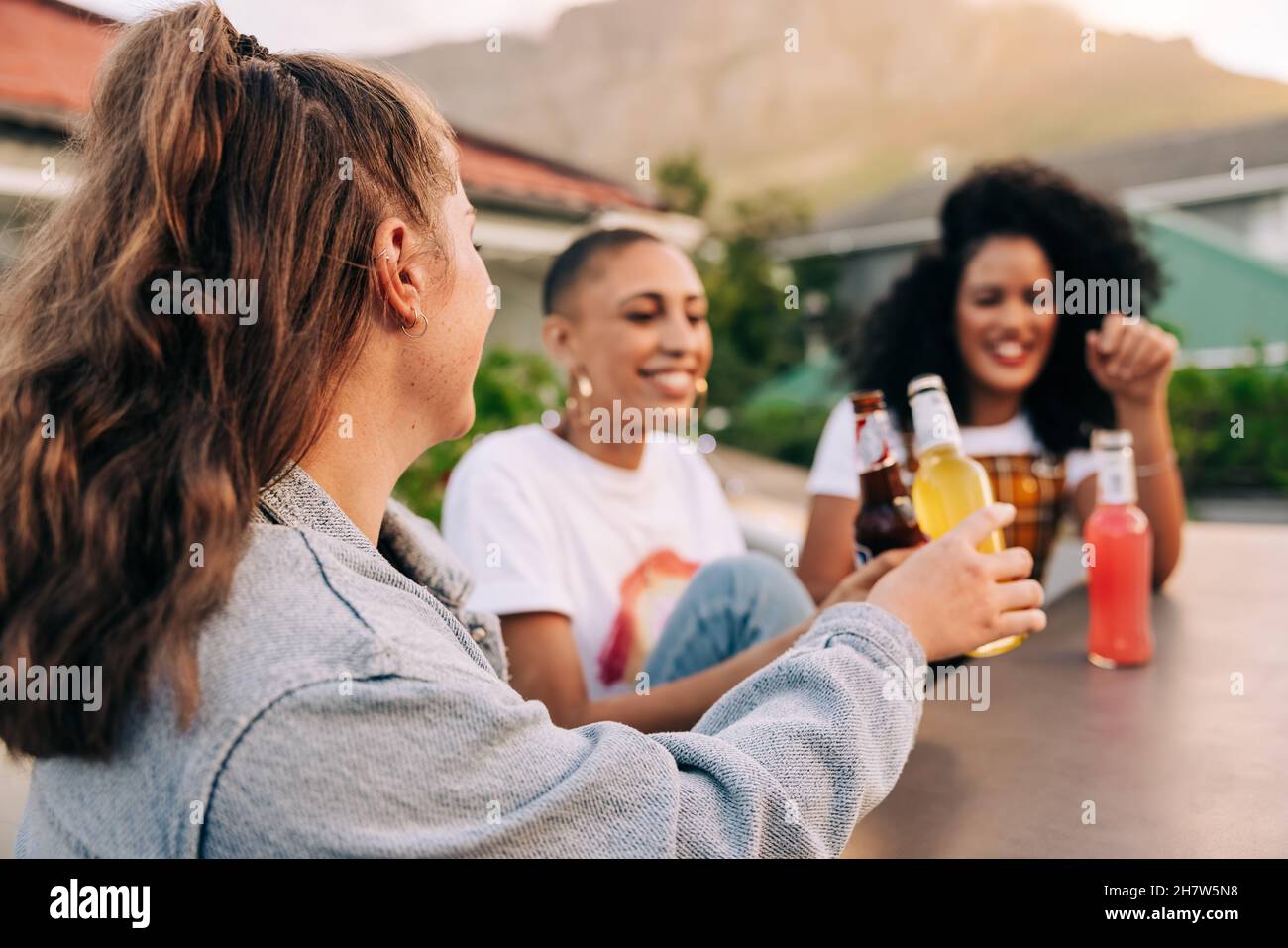 Tostar a la amistad. Feliz mujer joven haciendo un brindis con sus amigos en un tejado. Grupo de tres alegres amigas disfrutando de una cerveza fría Foto de stock