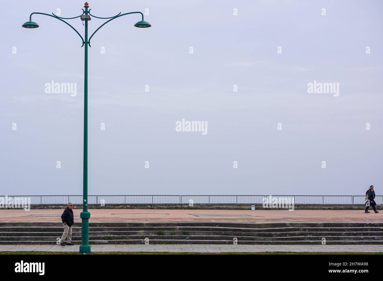 Vista desde la calle en el paseo marítimo de Lido, isla en la laguna de Venecia. Una pareja va a dar un paseo. Un hombre está parado por una lámpara de calle de doble iluminación. Foto de stock