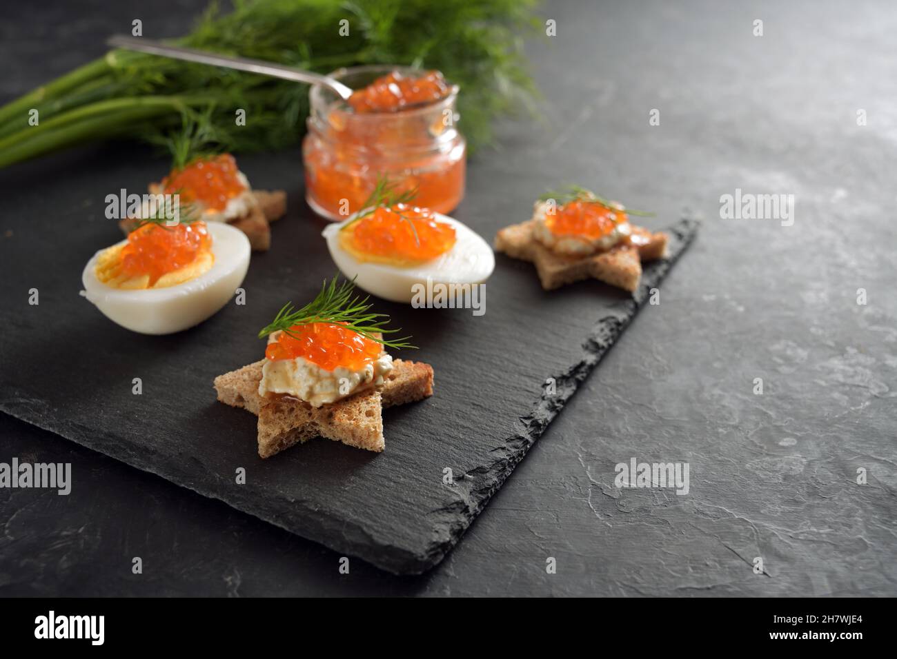 Caviar rojo en huevos a la mitad y en canapés tostados en forma de estrella con eneldo, preparación para un buffet festivo de Navidad en un plato de pizarra oscuro, copia grande Foto de stock