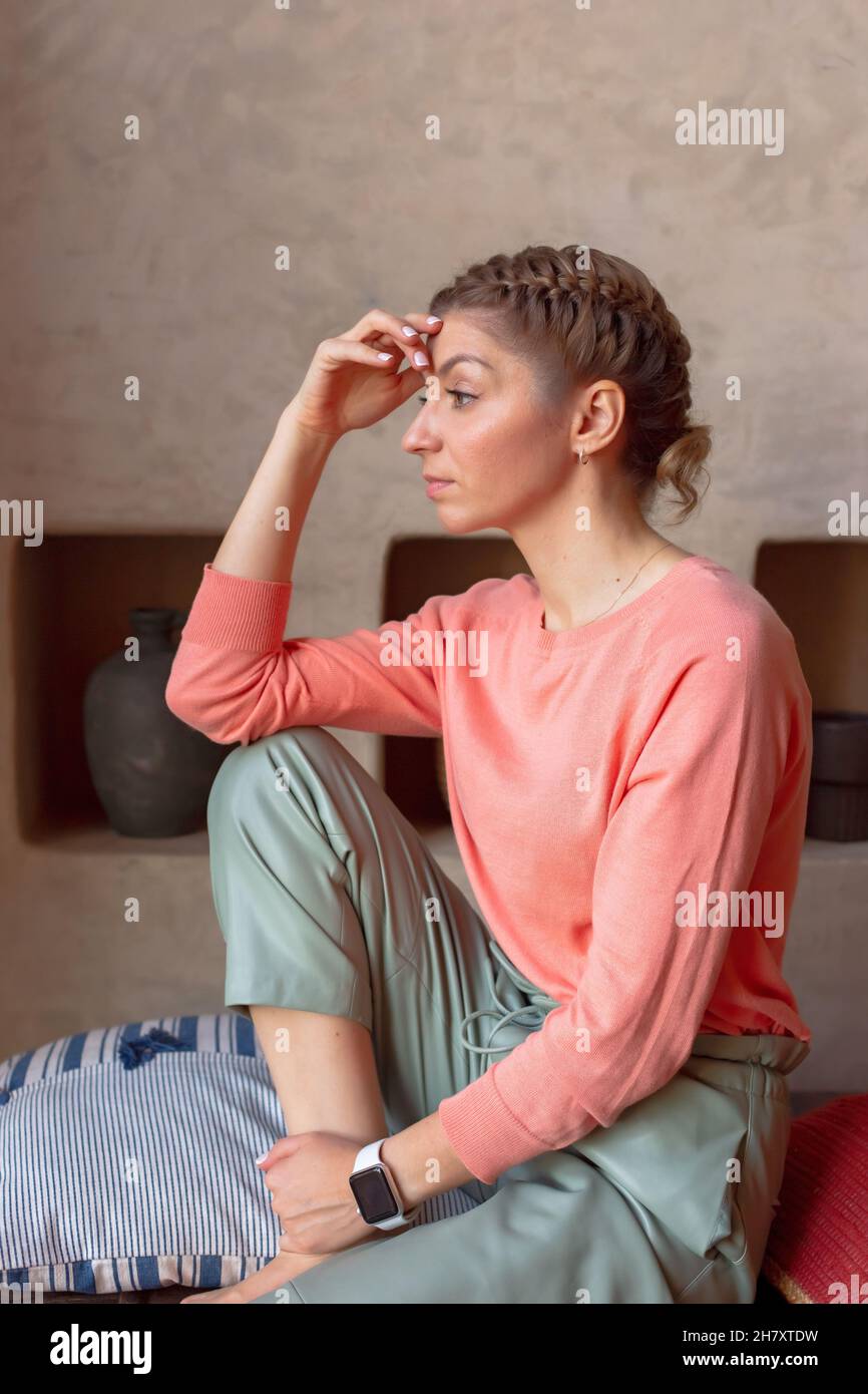 hermosa mujer pensante en sudadera de coral y pantalones grises está sentada en la habitación Foto de stock