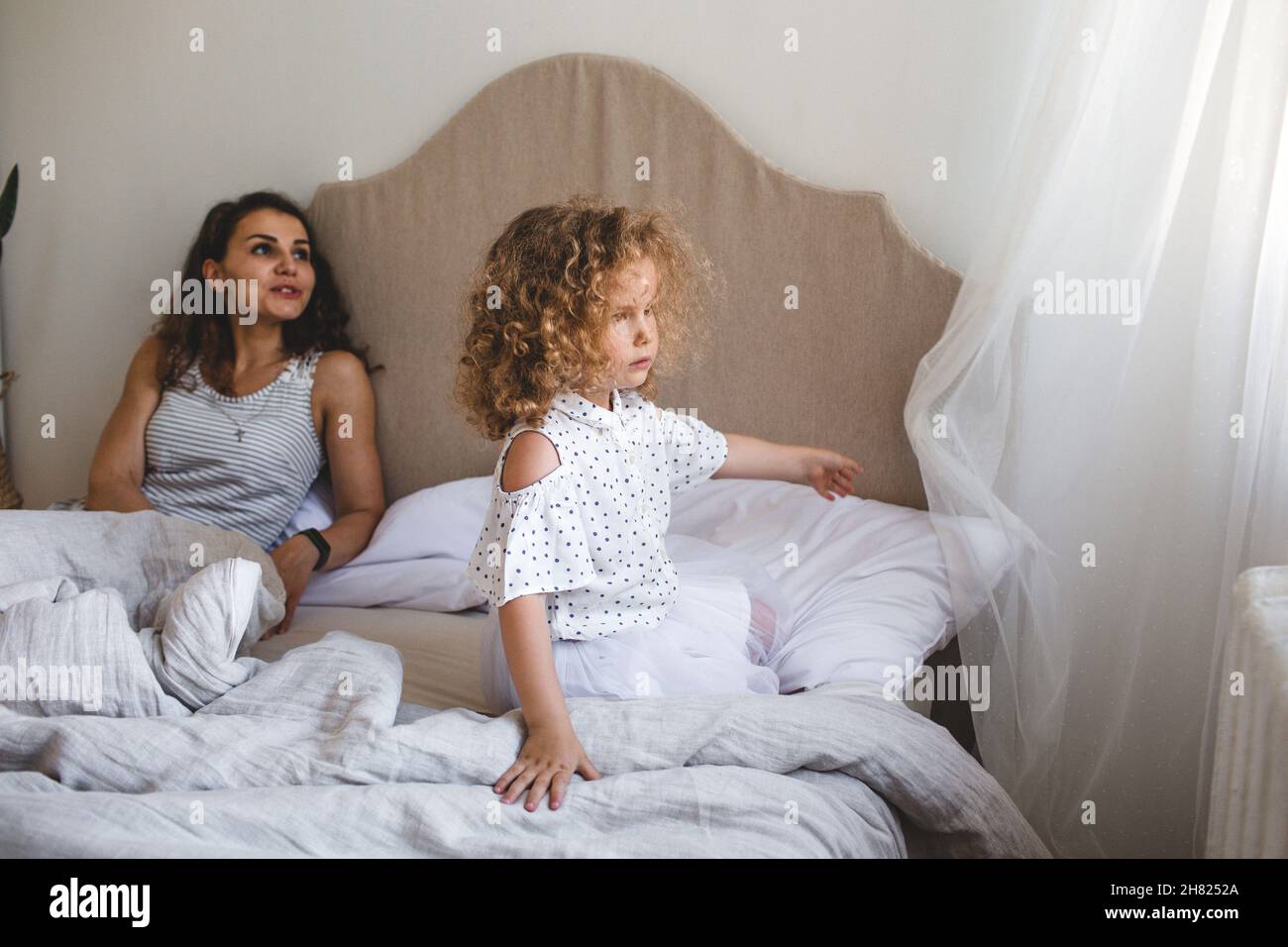 una niña está sentada en una cama grande con su madre mirando por la ventana. Foto de stock