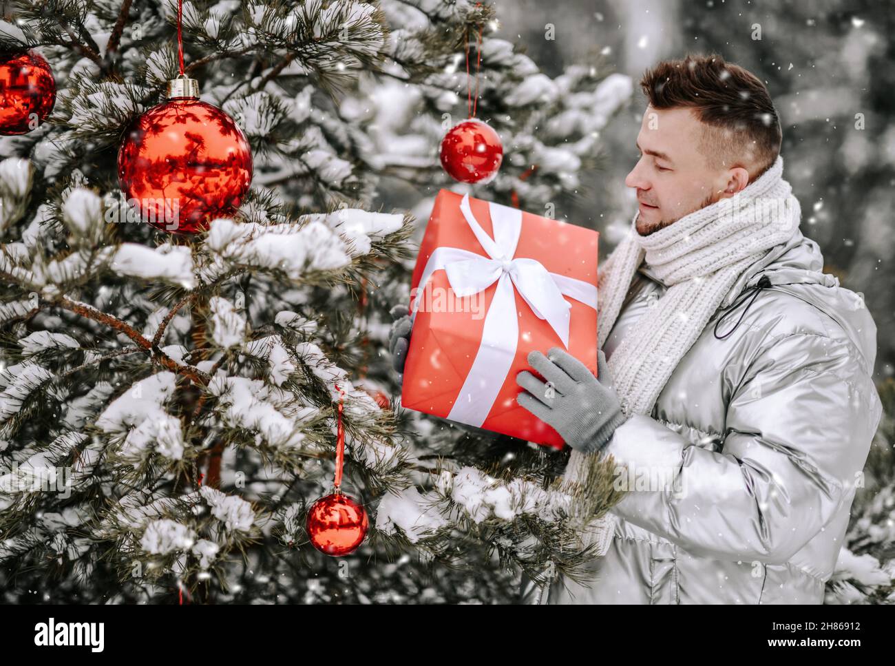Hombre joven con una elegante chaqueta plateada cerca del árbol de Navidad decorado y sosteniendo la caja de regalo de vacaciones al aire libre Foto de stock