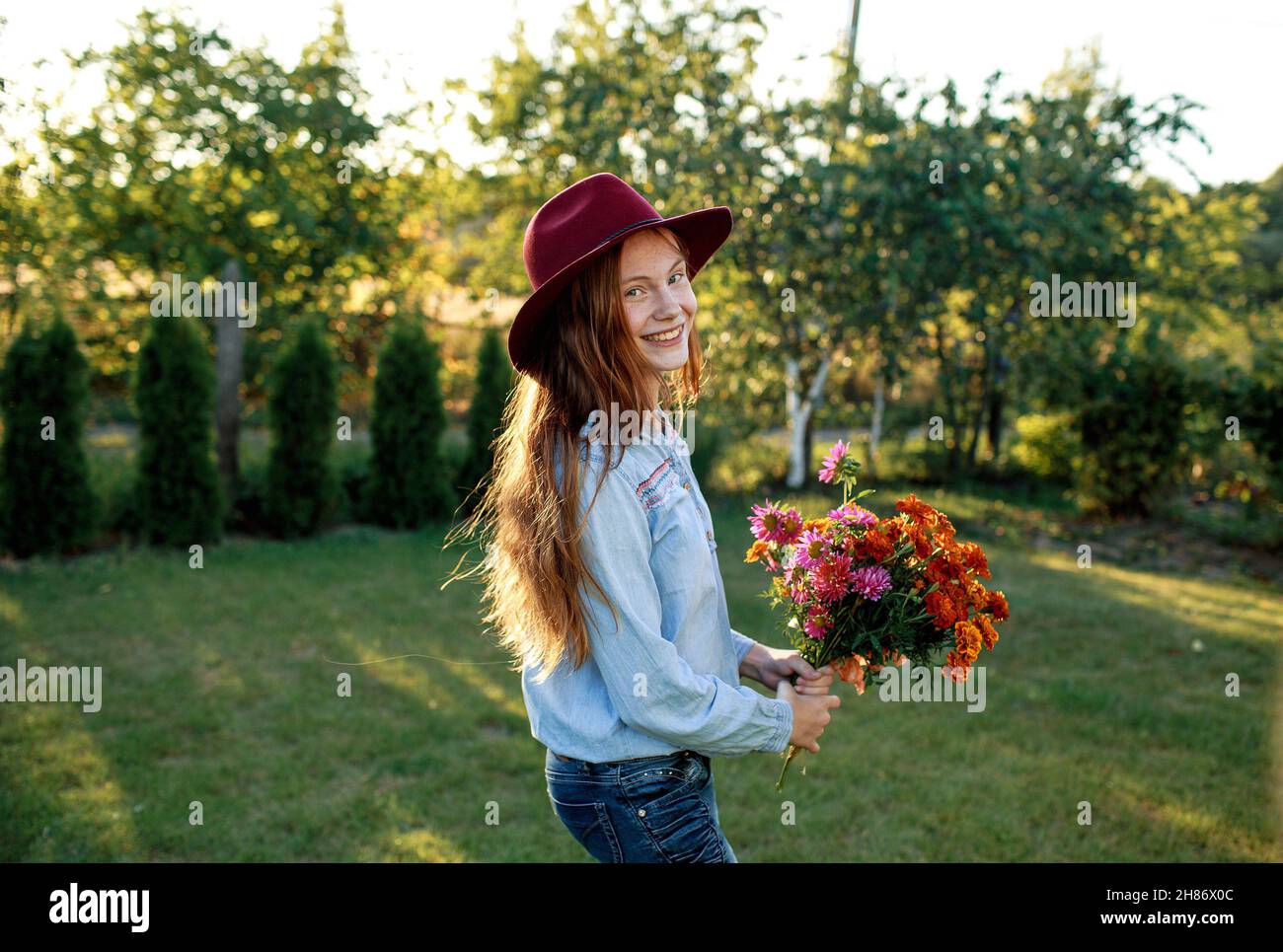 una adolescente de pelo rojo en un sombrero rojo baila en el césped con un ramo de flores Foto de stock