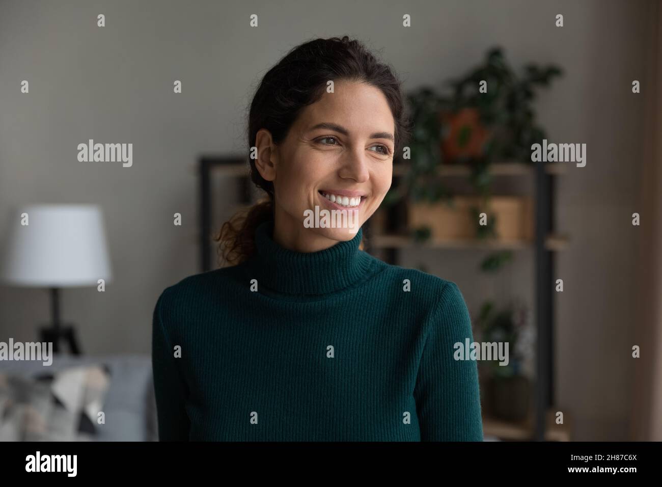 Sonriente mujer hispana sin preocupaciones de pie en la sala de estar mirando a un lado Foto de stock
