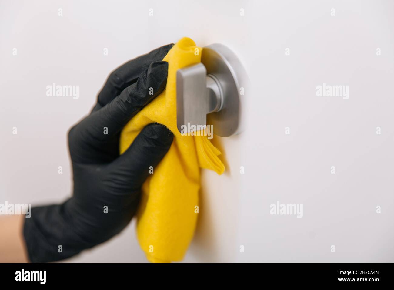 Limpieza del tirador de la puerta con un paño amarillo en guantes negros. Mano de mujer usando toalla para la limpieza. Desinfección en hospitales y espacios públicos contra corona Foto de stock