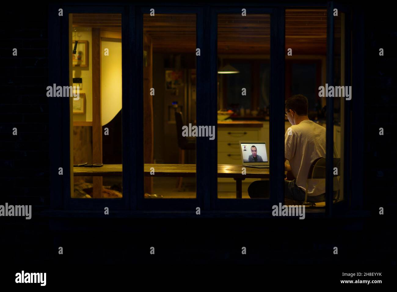 Un hombre que trabaja tarde en videoconferencias con un compañero de trabajo en el ordenador portátil de la cocina Foto de stock