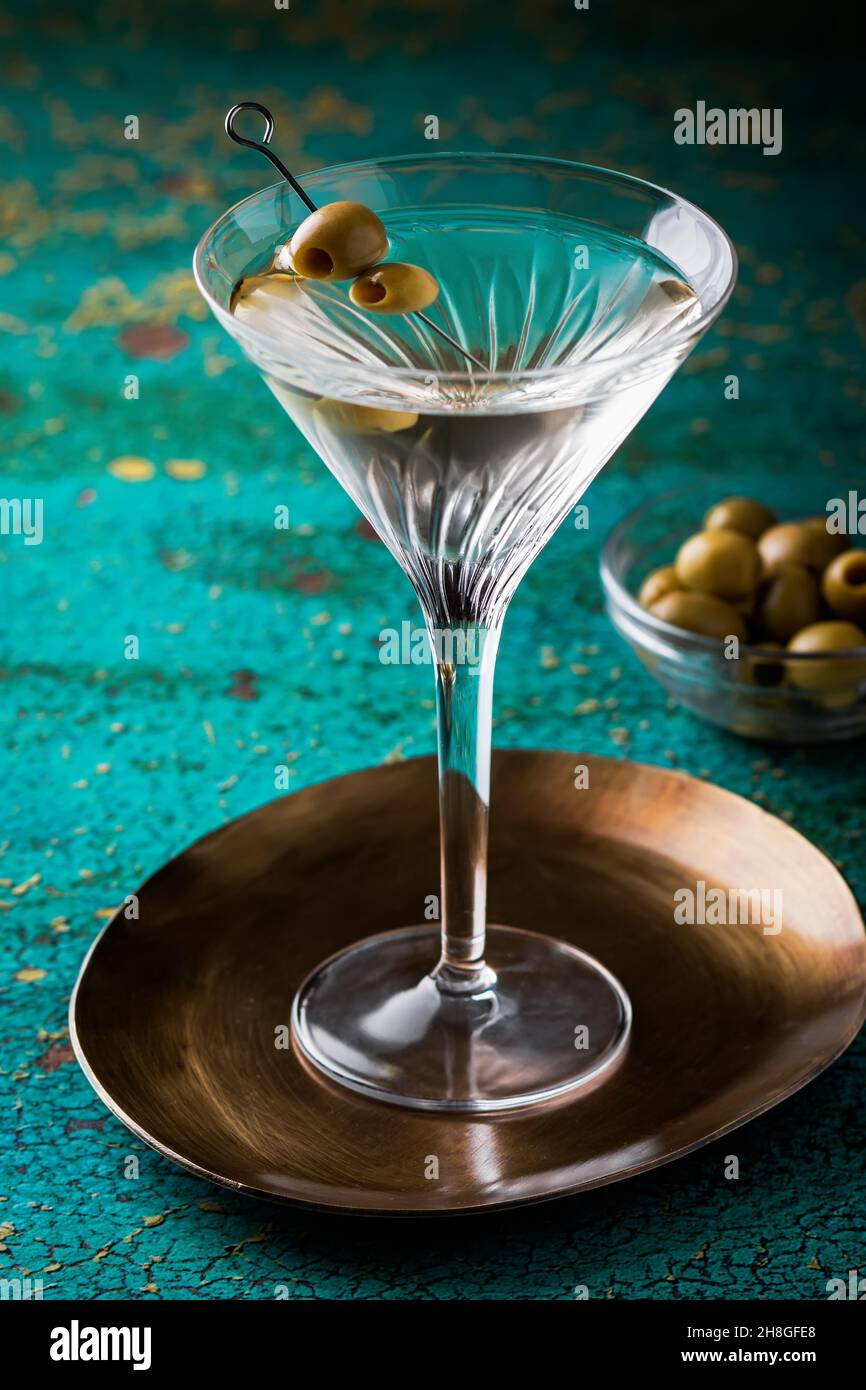 Cóctel martini sucio cierre en contraluz con aceitunas, adornar en mesa texturizada verde cierre con plato con aceitunas de fondo Foto de stock