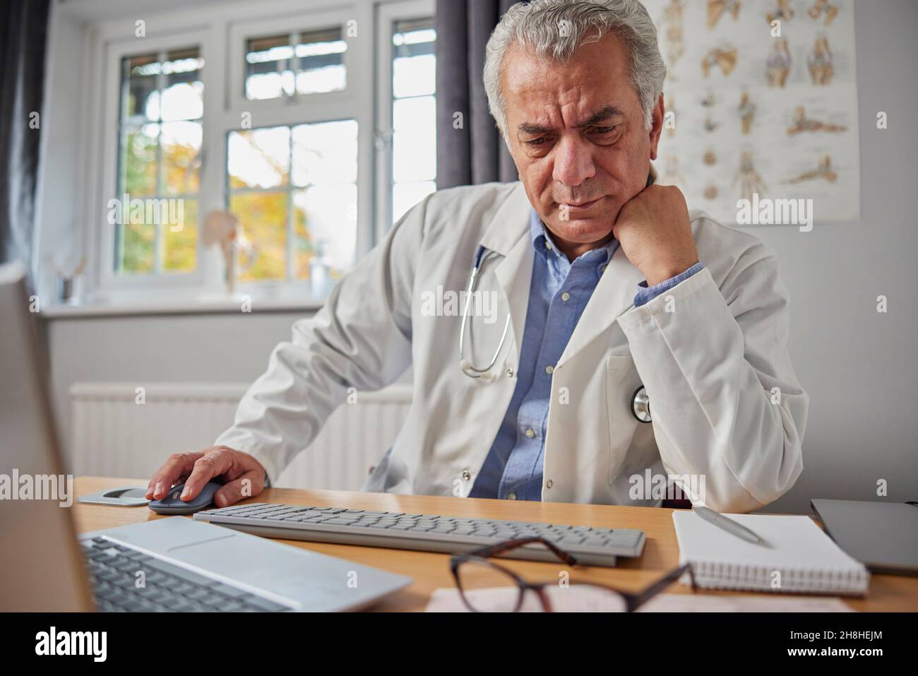 El GP Macho maduro estresado y trabajado en exceso usando el abrigo blanco en el escritorio en la oficina de los doctores Foto de stock