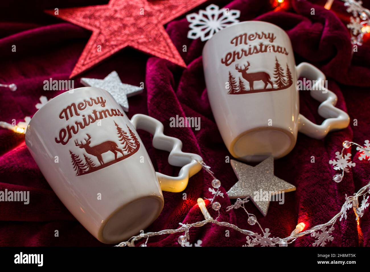 Dos tazas de Feliz Navidad en manta roja con luces y decoraciones Foto de stock