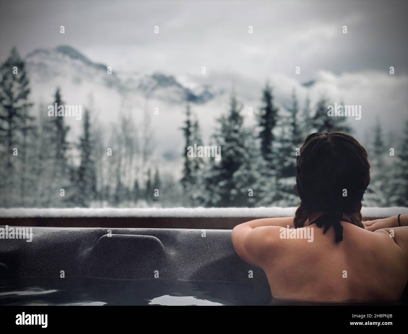 Mujer en la bañera de hidromasaje mirando a la vista de la montaña en BC Canadá, en el invierno Foto de stock