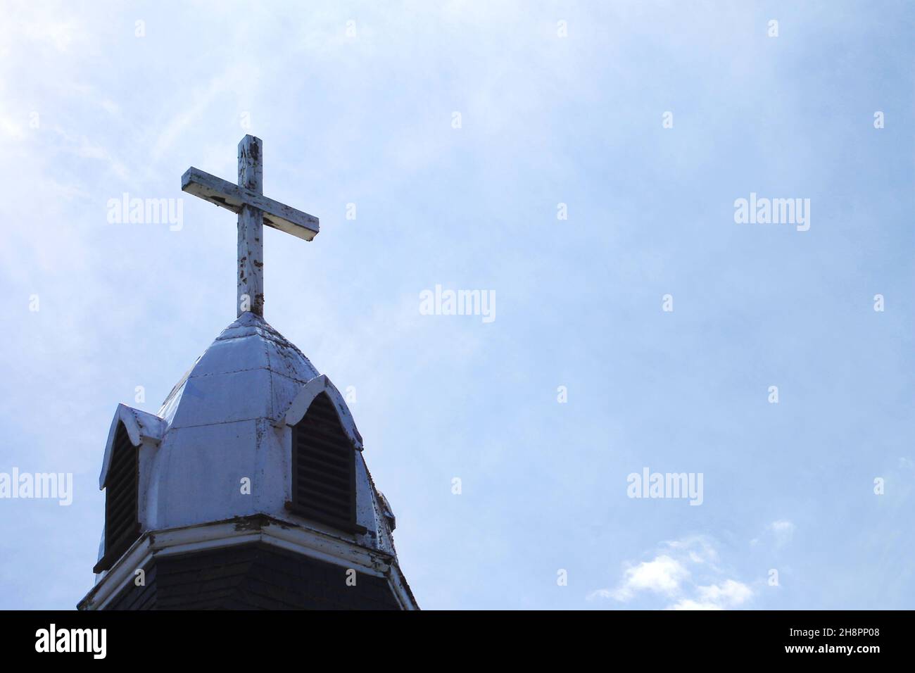 Primer plano de una cruz en la parte superior de un campanario de la iglesia. Cielo azul con nubes blancas y con aguas de fondo. Foto de stock