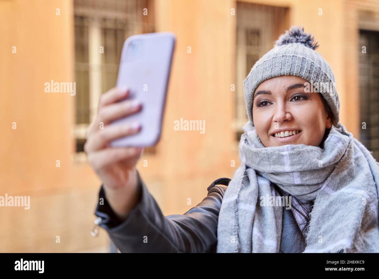 Mujer joven con estilo en ropa exterior sonriendo y tomando selfie mientras que parada en la calle de la ciudad en día frío Foto de stock
