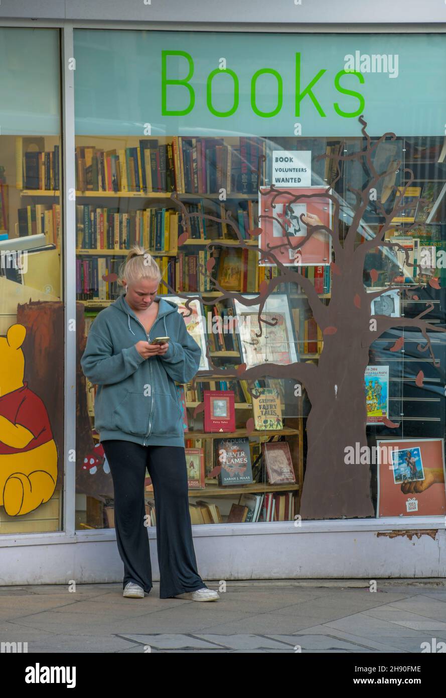 mujer joven que usa un teléfono móvil smartphone o dispositivo fuera de la ventana de una librería de oxfam, mujer leyendo mensajes telefónicos con la librería detrás Foto de stock