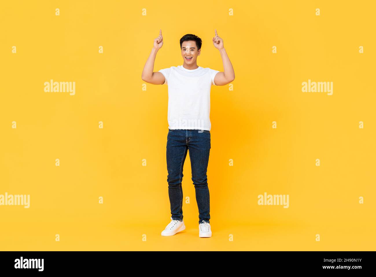 Retrato de cuerpo entero de hombre asiático sonriente y guapo apuntando dos dedos hacia arriba, aislado sobre fondo amarillo de estudio Foto de stock