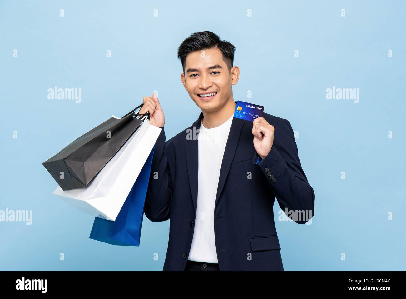 Joven hombre asiático guapo sosteniendo bolsas de compras y tarjeta de edición en un fondo aislado de color azul claro Foto de stock