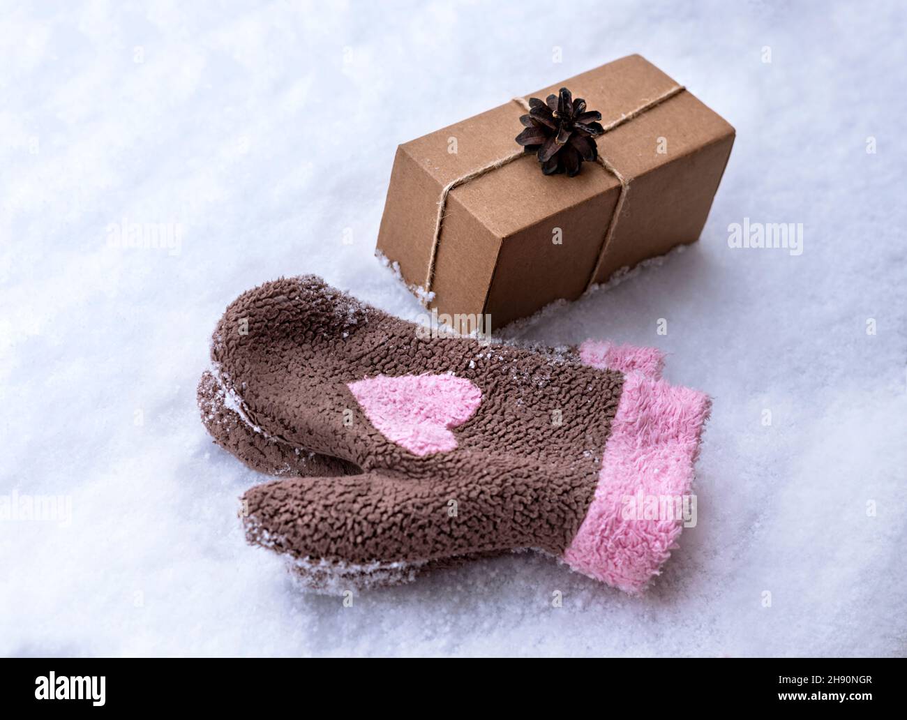 Regalo en una caja de artesanía decorada con cono de pino sobre nieve blanca y manoplas con corazón rosa, eco amistoso concepto Feliz Navidad y Año Nuevo Foto de stock