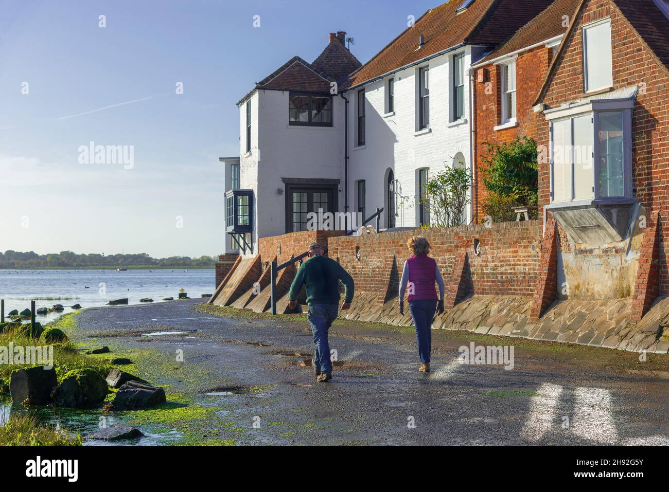 Pareja caminando sobre la marea caída a lo largo de la carretera de la costa en el pintoresco pueblo de Bosham, Chichester Harbor, nr.Chichester, West Sussex, Inglaterra Foto de stock