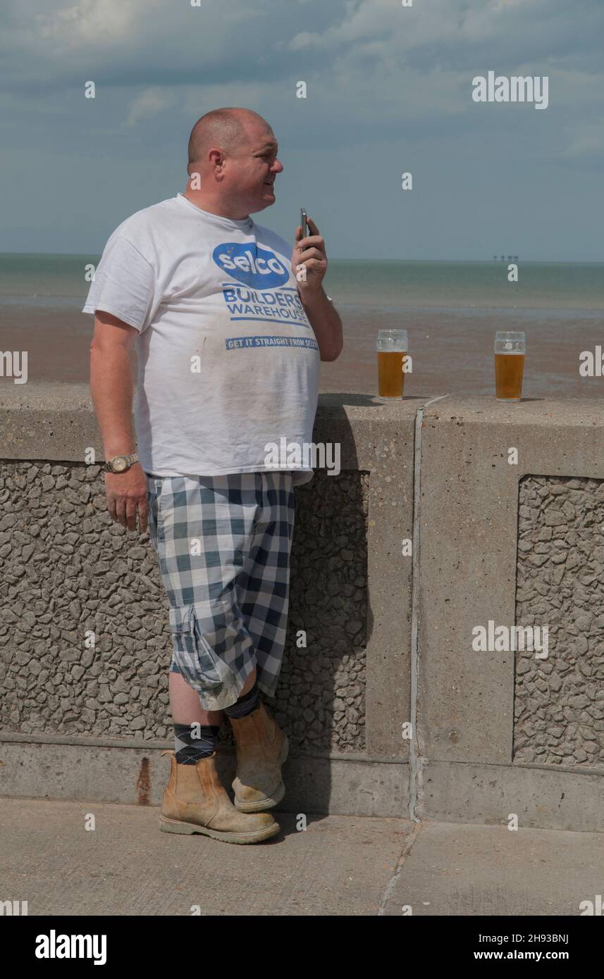 Hombre de clase trabajadora con iphone en su mano, en un fin de semana de vacaciones de verano. Pared de mar fuera del pub. Leysdown on Sea, Isla de Sheppey, Kent, Reino Unido 2014 2010s HOMER SYKES Foto de stock