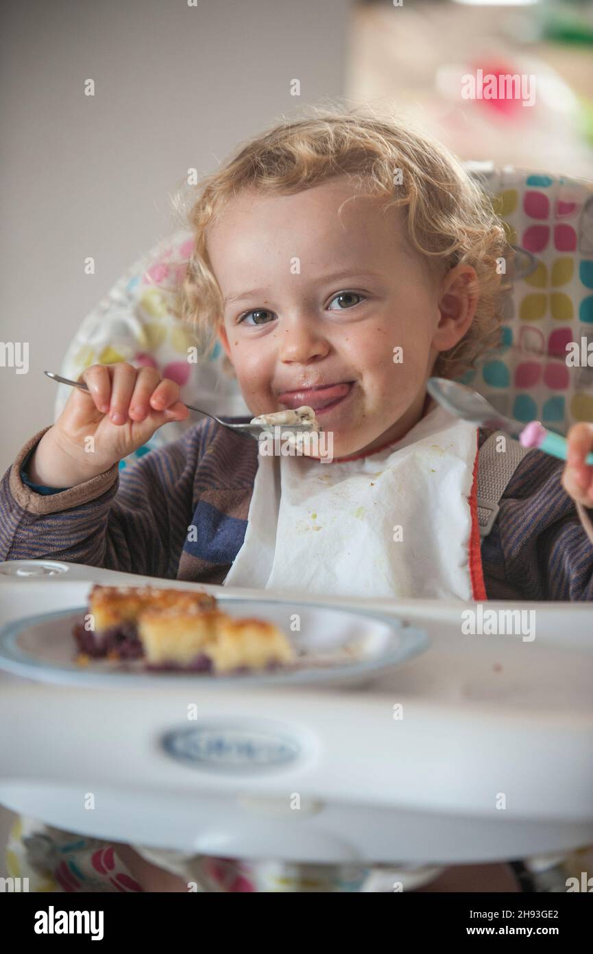 Una niña de 2 años come un poco de pastel y helado como aperitivo por la tarde en una trona. Foto de stock