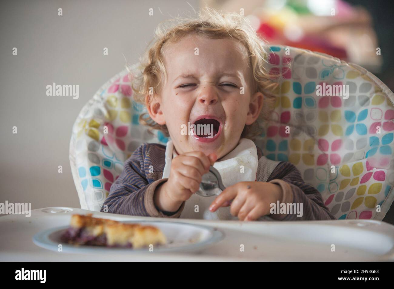 Una niña de 2 años llora mientras se sienta en una silla alta con algo de pastel como merienda por la tarde Foto de stock