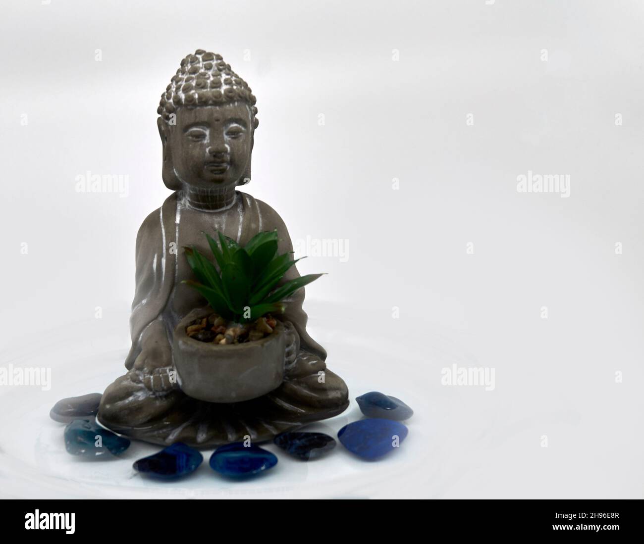 Un pequeño Buda gris con un suculento rodeado de siete piedras azules semi-preciosas en el agua. Fondo blanco. Foto de stock