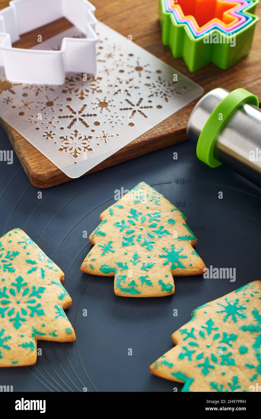 Galletas con forma de árbol de Navidad con patrón de copos de nieve, plantilla, cortadores de galletas, pasador rodante, tabla de cortar en una alfombrilla para hornear Foto de stock