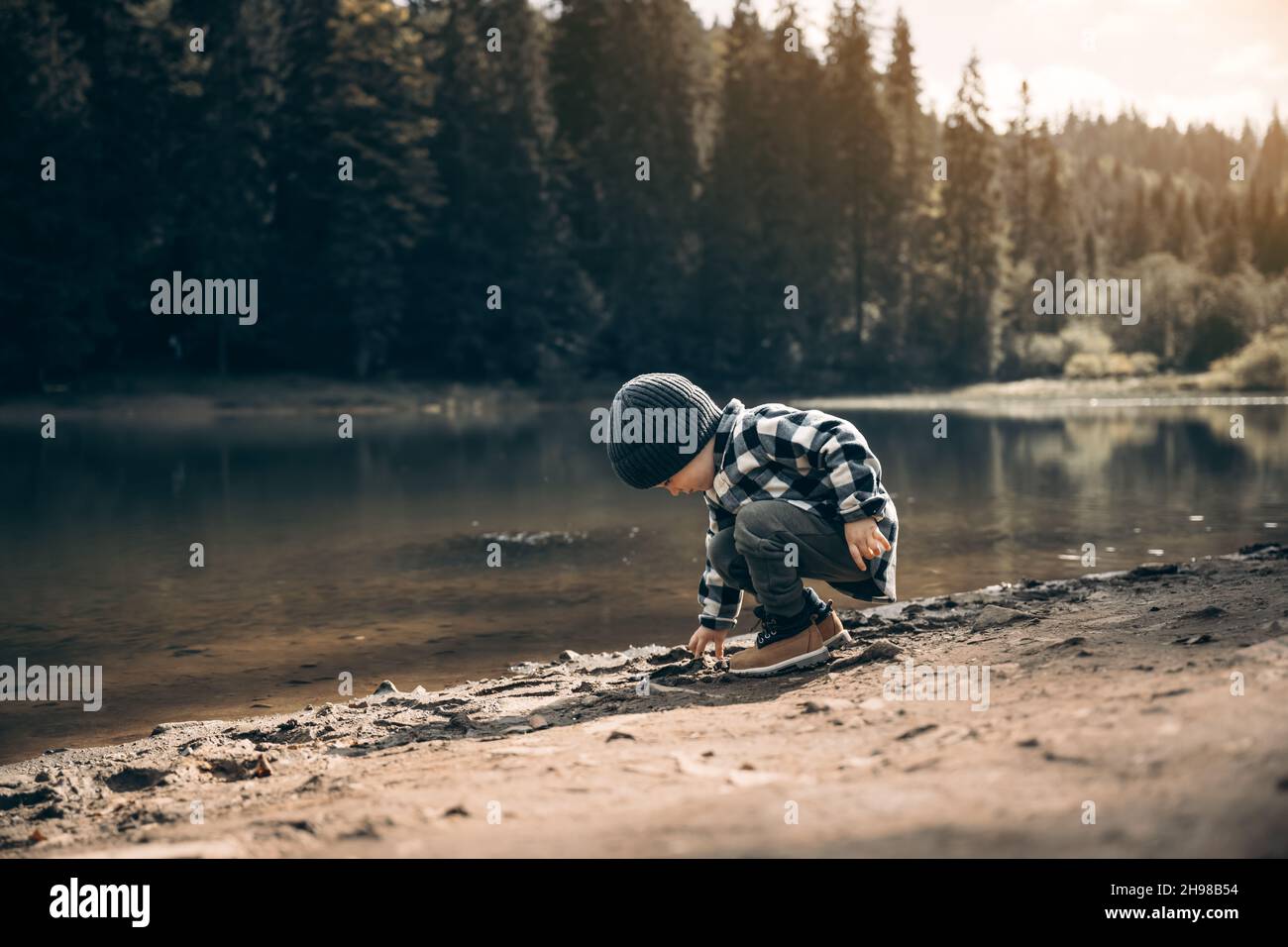 Niño pequeño en una camisa de cuadros y sombrero gris en la costa del lago del bosque. Infancia con concepto amante de la naturaleza Foto de stock