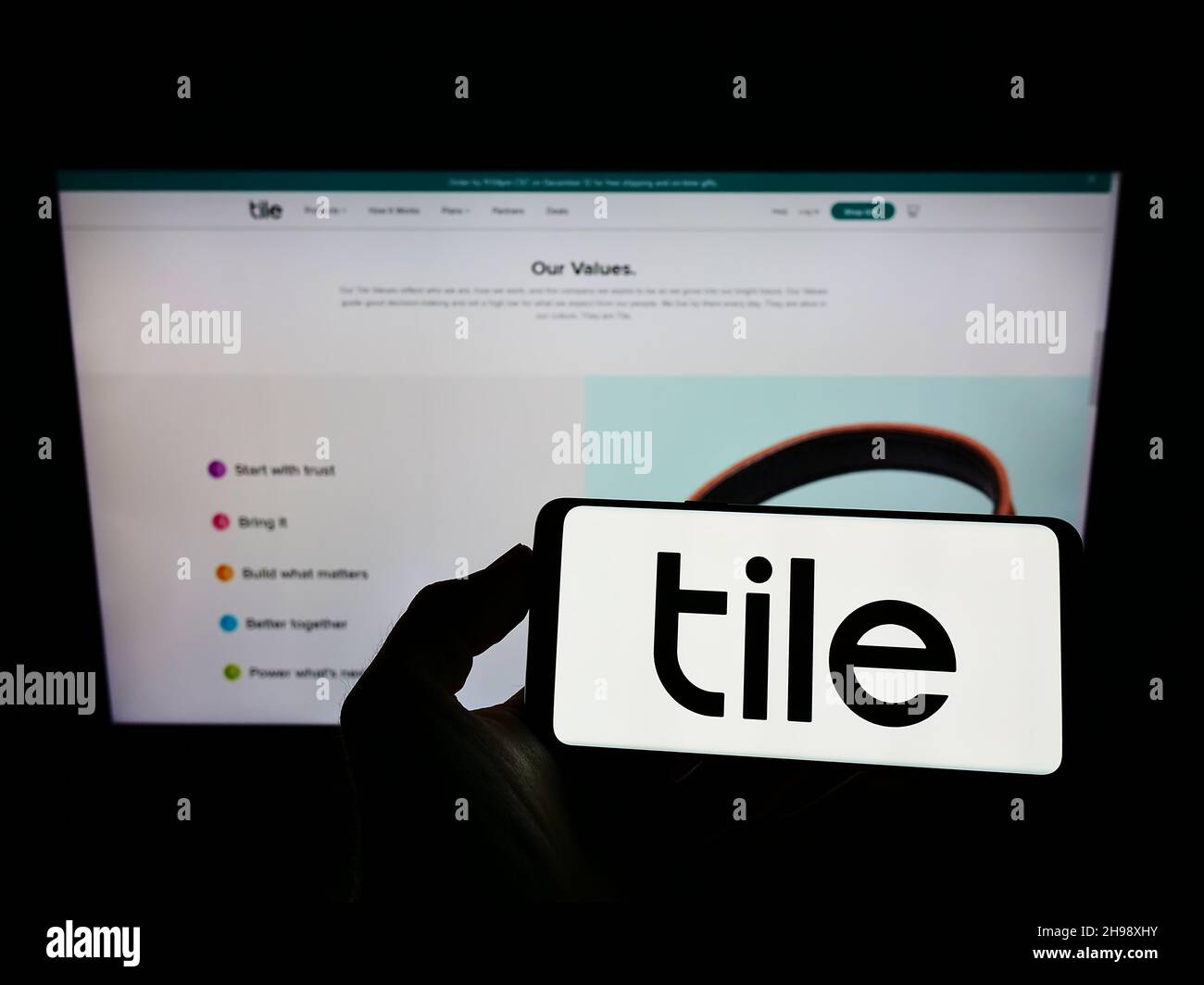 Persona que sostiene el teléfono móvil con el logotipo de la empresa estadounidense de electrónica de consumo Tile Inc. En la pantalla frente a la página web. Enfoque en la pantalla del teléfono. Foto de stock