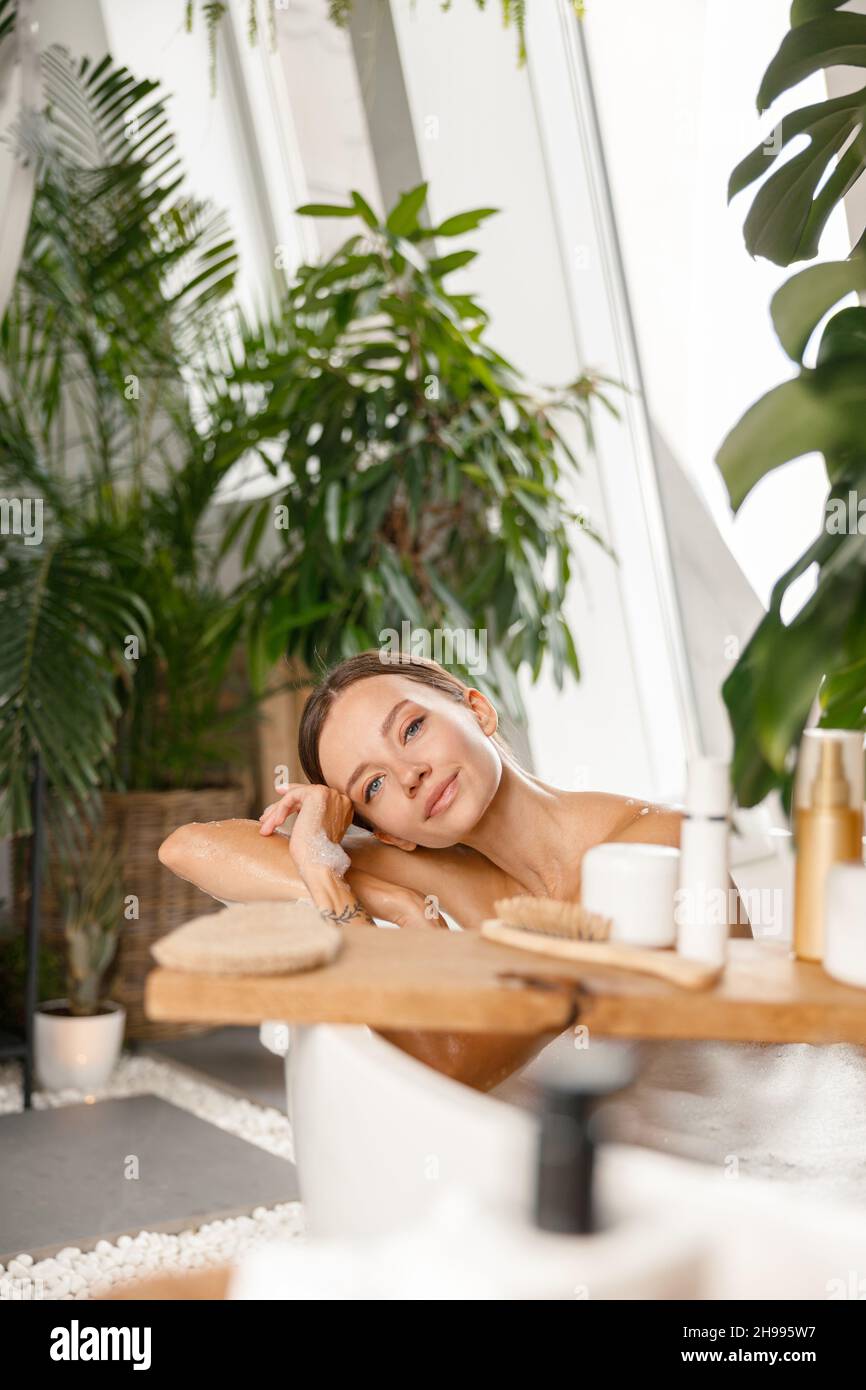 Una mujer joven y tranquila mirando la cámara mientras se toma un baño de burbujas en un spa tropical Foto de stock