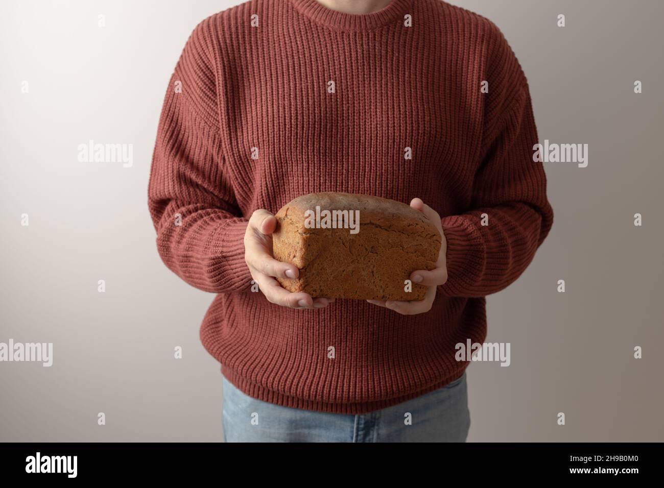 Un hombre en un suéter tiene una barra de pan de centeno casero. Vista frontal. Foto de stock