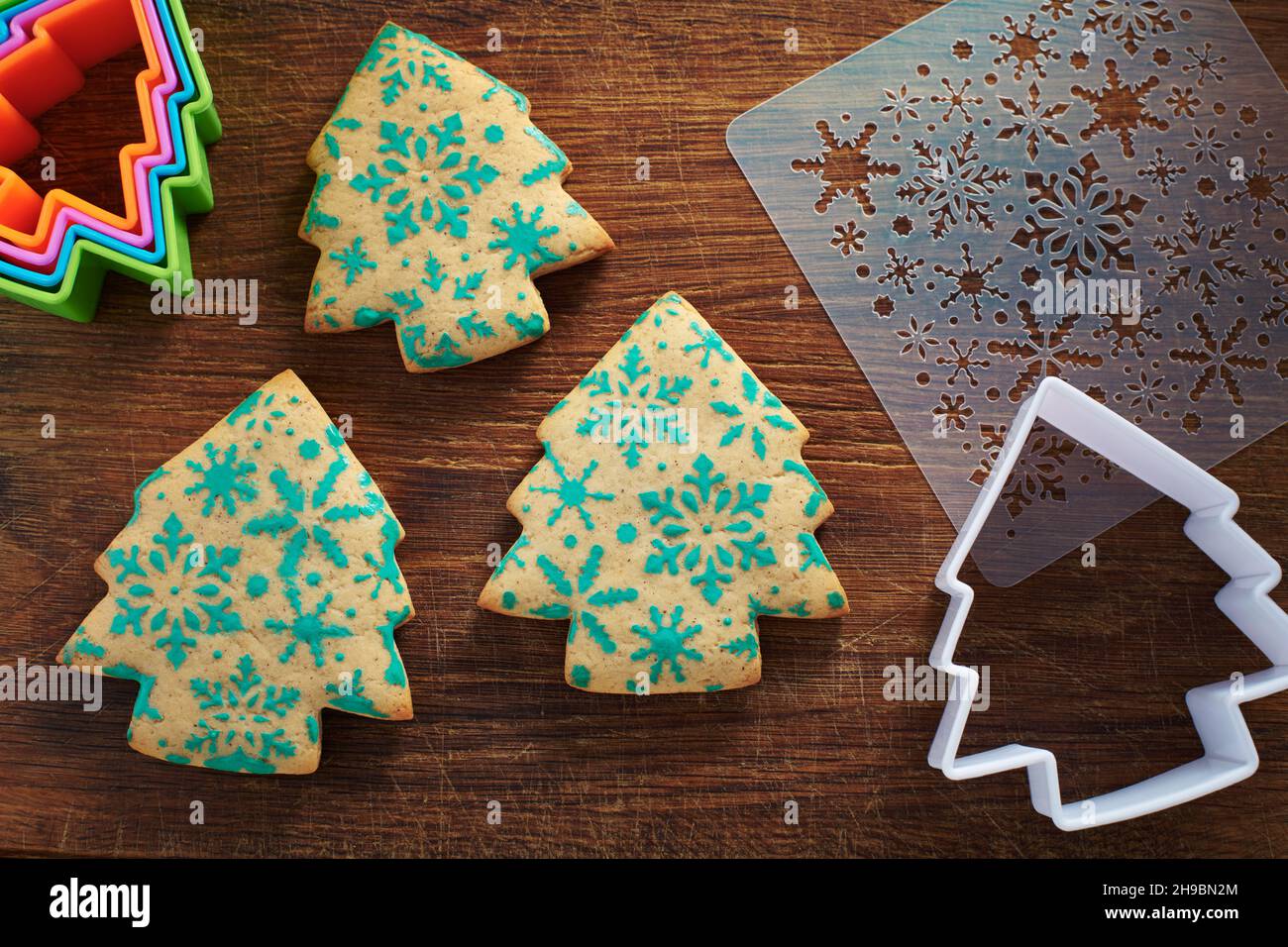 Galletas con forma de árbol de Navidad con patrón de copos de nieve, cortadores de galgas y galletas en una tabla de cortar de madera Foto de stock